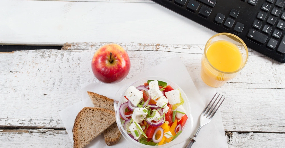 Auf einem weißen Holztisch befinden sich ein bunt gemischter Salat in einer Schüssel, ein Glas Orangensaft, ein Apfel, zwei Scheiben Brot und eine Gabel neben einer Computertastatur.