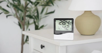 Auf weißem Beistelltisch mit Tischleuchte im Hintergrund und einer Zimmerpflanze links davon steht ein Hygrometer, das eine Raumtemperatur von 28,4 Grad und eine Luftfeuchte von 58 % anzeigt.