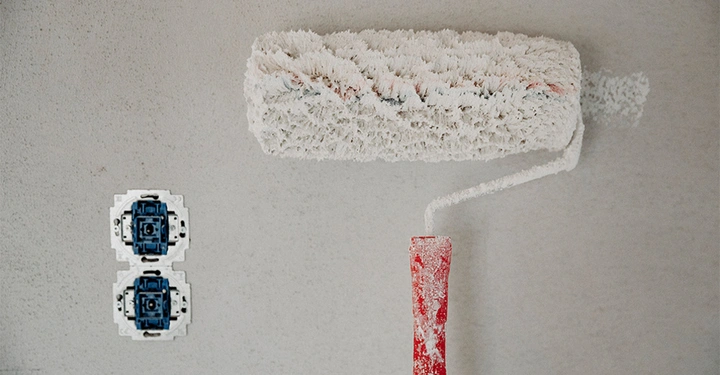 Ein Farbroller mit weißer Farbe wird an einer Wand verwendet. Daneben sind zwei unmontierte Steckdosen mit blauen Komponenten sichtbar.