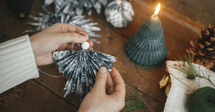 Hände halten eine Papierdekoration ein angezündetes Baumkerzenlicht und eine Tannenzapfendekoration auf einem hölzernen Tisch mit grünem Garn und Kiefernnadeln im Umfeld