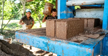 Schlammziegel werden von einer blauen Maschine geformt während zwei Arbeiter im Hintergrund in einer grünen Außenumgebung Materialien hinzufügen