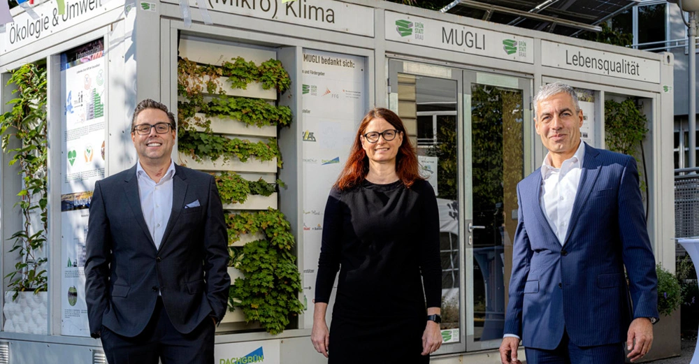 Drei Personen stehen vor einem modularen Gebäude mit vertikaler Begrünung und Schildern Mügli Mikroklima Ökologie & Umwelt Lebensqualität umgeben von Pflanzen und Bäumen. Text auf der Tafel: "MÜGLI bedankt sich: ...".