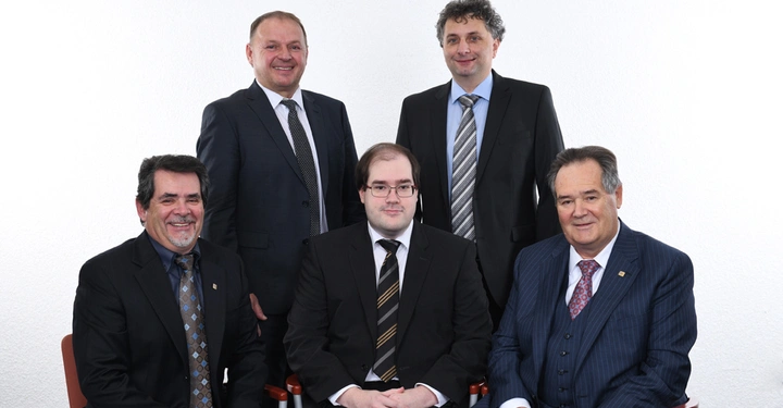 Fünf Männer in Anzügen sitzen und stehen in einem Raum mit weißer Wand. Drei Männer sitzen in der vorderen Reihe, während zwei hinten stehen. Alle schauen ernst oder lächeln leicht.