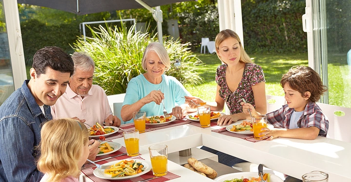 Sechs Personen essen am Tisch leichtes Mittagessen im hellen Raum draußen im Gartenoval.