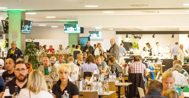 Galerie-Bild 3: Menschen essen und unterhalten sich in einem vollen Restaurant mit mehreren Fernsehern und stilvollem Interieur. Servicemitarbeiter stehen hinter einem Buffet und servieren Essen und Getränke. Text: "RAPID".