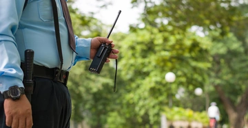 Der Wachmann trägt eine blaue Uniform und hält ein Funkgerät in der Hand. Im Hintergrund befindet sich ein parkähnlicher Außenbereich mit Bäumen und einem Fußgänger auf einem Weg.