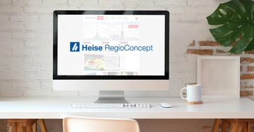 Ein Computermonitor zeigt das Logo von Heise RegioConcept auf einem Schreibtisch mit einer Kaffeetasse einer Pflanze an einer Ziegelwand.
