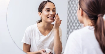 Frau trägt eine weiße Bluse trägt Gesichtscreme auf steht vor einem runden Spiegel in einem Badezimmer mit gemusterten Fliesen im Hintergrund
