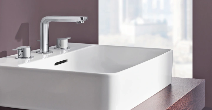 Ein quadratisches weißes Waschbecken mit einem modernen Chrom-Wasserhahn und zwei Drehknöpfen auf einer Holzoberfläche vor einer lila gestrichenen Wand und einem Fenster mit verschwommener Stadtansicht.