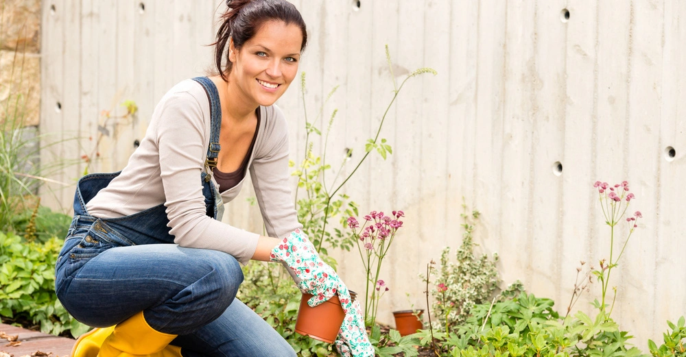 Junge Frau mit braunen, zurückbebundenen Haaren in Jeanslatzhose und Gartenhandschuhen setzt Blumen aus Topf in Erde. Lächelt freundlich in die Kamera