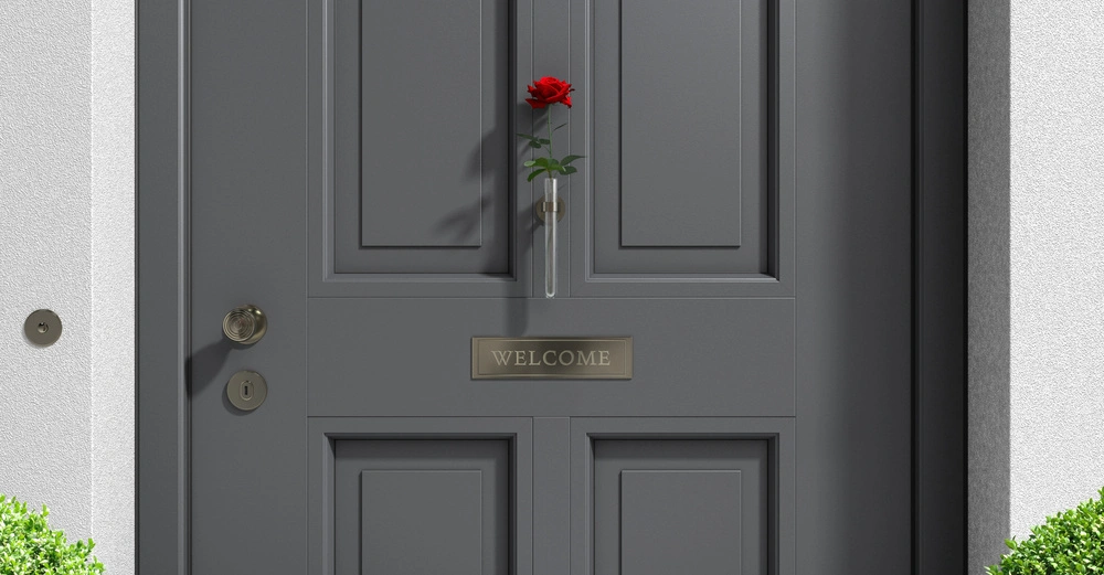 Graue Haustür hält rote Rose in einem kleinen Glasbehälter; Kontext ist ein Eingang mit zwei grünen Pflanzen auf beiden Seiten. Beschriftung WELCOME auf einem goldenen Schild.