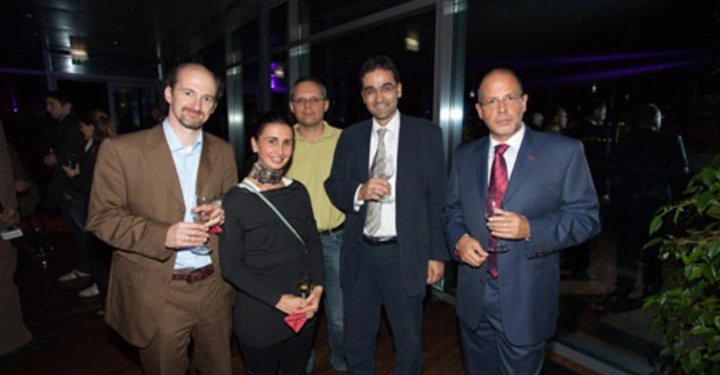 Galerie-Bild 73: Menschen stehen und lächeln in Abendkleidung, halten Getränke in den Händen, in einem modernen Raum mit Glaswänden und Pflanzen, während andere Personen im Hintergrund an einem gesellschaftlichen Event teilnehmen.