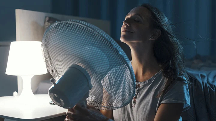 Frau kühlt sich nachts während einer Hitzewelle mit einem Ventilator im dunklen Schlafzimmer an.