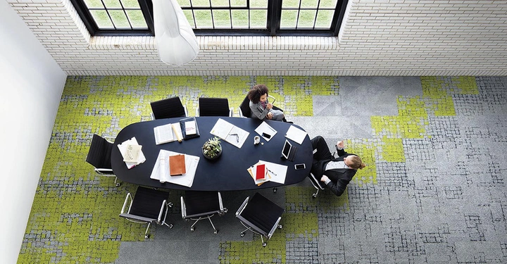 Galerie-Bild 7: Zwei Personen sitzen an einem ovalen Tisch und unterhalten sich in einem modernen Raum mit großen Fenstern und gemustertem Teppichboden der Tisch ist mit Laptops Büchern und Papieren bedeckt