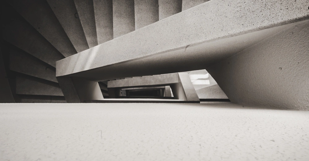Treppenstufen verlaufen spiralförmig nach oben in einem modernen Betongebäude. Schmale Betonwände bilden geometrische Muster und betonen die Perspektive. Einfallendes Licht erzeugt Schatten und Kontraste in der Szene.