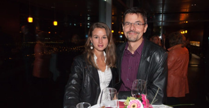 Galerie-Bild 77: Zwei Personen sitzen an einem gedeckten Tisch in einem schicken Restaurant trinken Wein und lächeln in die Kamera. Der Hintergrund zeigt ein abendliches Stadtpanorama durch große Fenster.