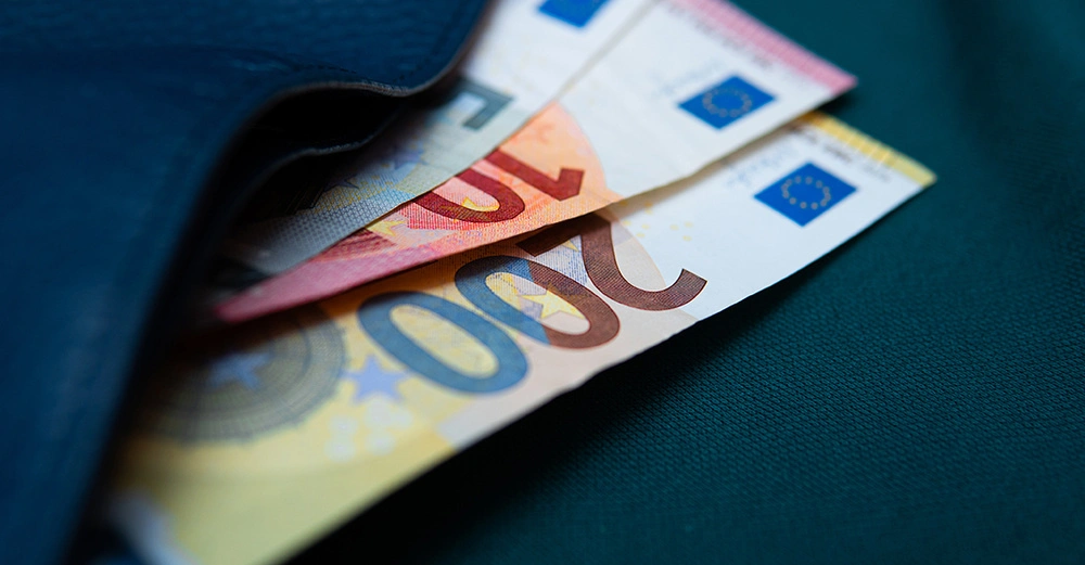 Banknoten in den Werten von 20 und 200 Euro ragen aus einer schwarzen Brieftasche auf einem dunkelgrünen Tuch heraus.