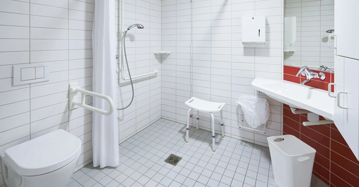 Toilette mit Haltegriffen und Duschvorhang in einem behindertengerechten weißen Badezimmer mit Sitzstuhl in der Dusche Waschbecken Spiegel und Mülleimer auf Fliesenboden.