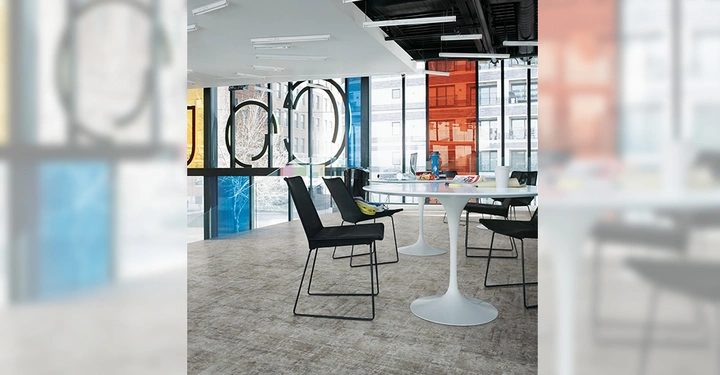 Galerie-Bild 4: Mehrere schwarze Stühle stehen um runde weiße Tische in einem modernen Büroraum. Tageslicht durch bunte Fenster fällt auf die helle Innenausstattung. Bürogebäude sind durch die Fenster sichtbar.