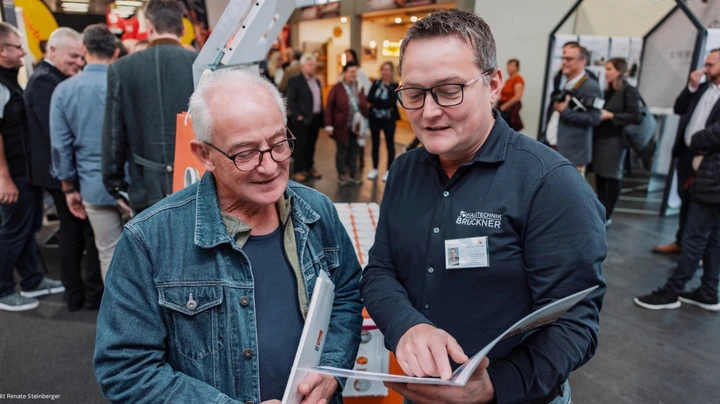 Galerie-Bild 4: Älterer Herr mit Brille blickt interessiert in ein Buch, in dem ein jüngerer Experte auf etwas zeigt.