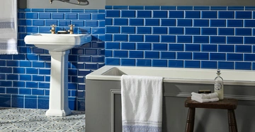 Waschbecken aus Porzellan mit Handtuch an Halterung. Badewanne mit aufgehängtem Handtuch. Holzhocker daneben mit Seife und Flasche. Blau-weiße Fliesen an Wand und Boden im Badezimmer.