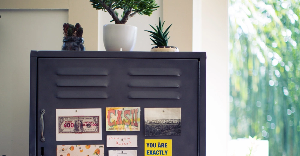 Ein grauer Stahlschrank steht mit mehreren Pflanzen darauf in einem hellen Raum. Auf dem Schrank sind verschiedene Notizen und Bilder angebracht. Text: CASH STAY CONNECTED YOU ARE EXACTLY WHERE YOU NEED TO BE