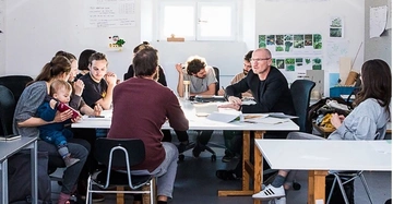 Professoren und Studierende sitzen locker diskutierend an großem Tisch in hellem Raum.