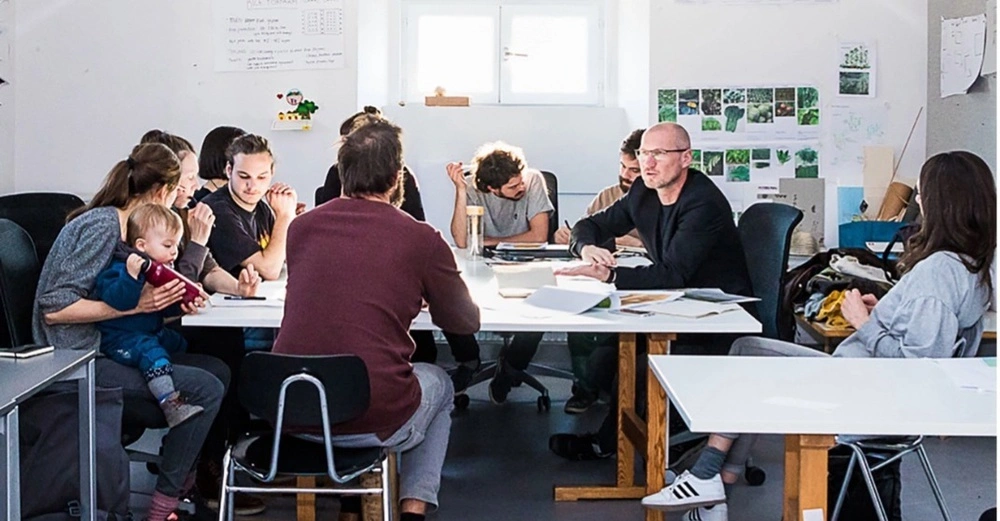 Professoren und Studierende sitzen locker diskutierend an großem Tisch in hellem Raum.