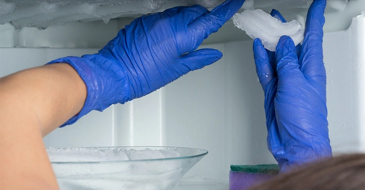 Hände in blauen Handschuhen entfernen Eisstücke im Inneren eines Gefrierschranks