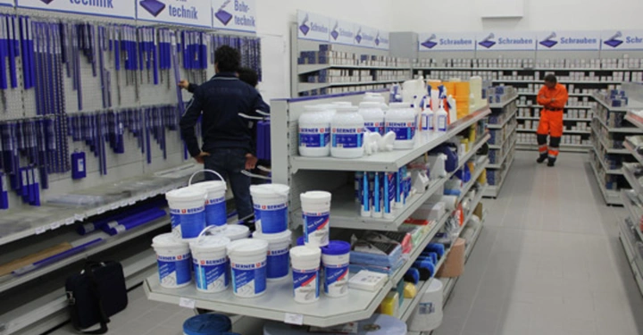 Galerie-Bild 9: Eine Person sortiert Bohrtechniken an einer Wand in einem Baumarkt ein eine andere Person in orangener Arbeitskleidung geht durch die Gänge es gibt viele Regale mit verschiedenen Baumarktartikeln.