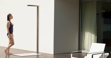 Frau in Badeanzug geht zu einer Außendusche vor einem modernen Gebäude mit weißem Sessel und großem Fenster.