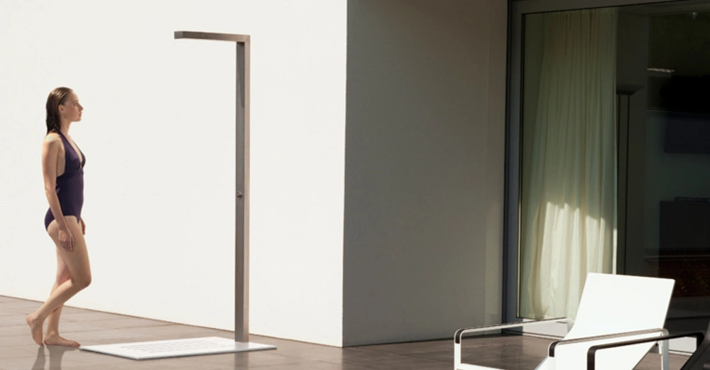 Frau in Badeanzug geht zu einer Außendusche vor einem modernen Gebäude mit weißem Sessel und großem Fenster.