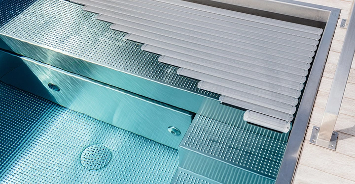 Ein teilweise aufgerollter, metallischer Poolabdeckungsmechanismus öffnet sich über einem blauen, rechteckigen Schwimmbecken, das von einem weißen Fliesenrand umgeben ist.