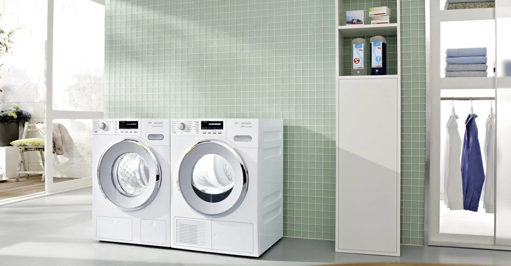 Zwei Waschmaschinen nebeneinander stehend in einem hellen Raum mit grünen Kachelwänden Regale mit Waschmittel und gefalteter Wäsche befinden sich rechts Waschbecken und Pflanzen sind auf der linken Seite
