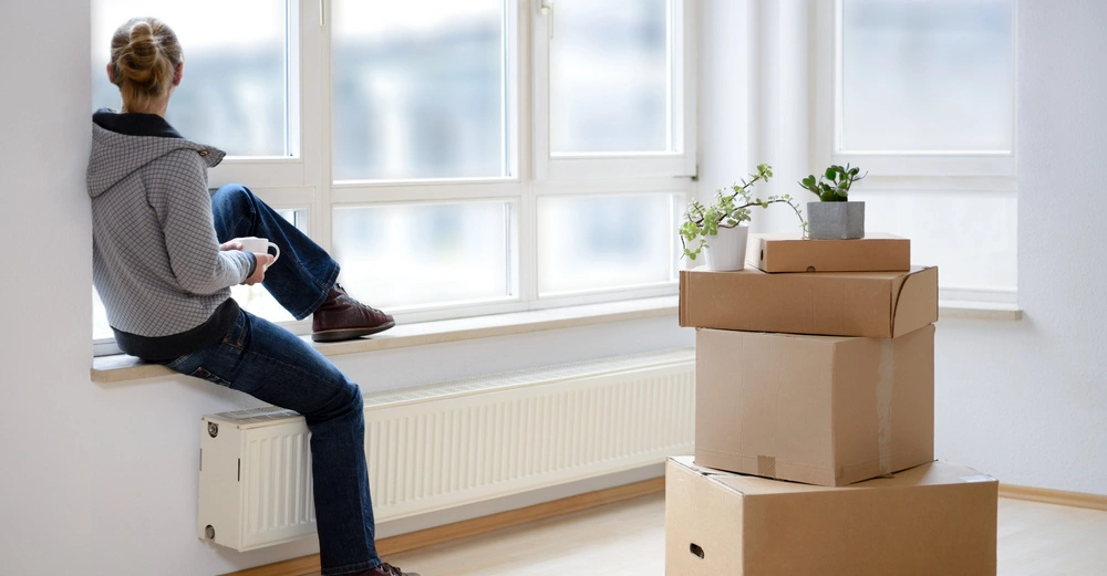 Eine sitzende Person trinkt aus einer Tasse und schaut aus dem Fenster in einem leeren Raum mit einem Stapel Umzugskartons und Zimmerpflanzen im Vordergrund.