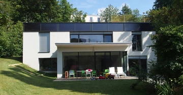 Ein modernes zweistöckiges Haus steht in einem gepflegten, grünen Garten. Auf der Terrasse vor dem Haus befinden sich bunte Stühle, Spielzeuge und Möbel. Bäume umgeben das Haus im Hintergrund.