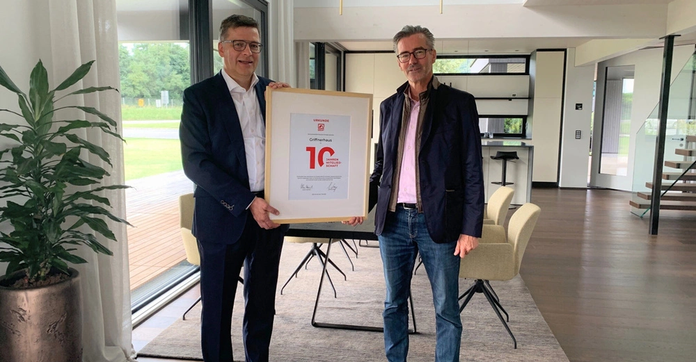 zwei Männer im Anzug stehen in hellem Büroraum und halten große Urkunde in der Hand. Übergabe von Lange an Niedersüß.