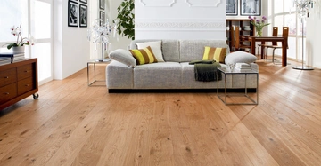 Graues Sofa mit grünen und gelben Kissen steht in einem hellen Wohnzimmer vor einem Tisch auf einem Holzfußboden; im Hintergrund ein Essbereich mit Holztisch und Stühlen sowie Wandbilder.