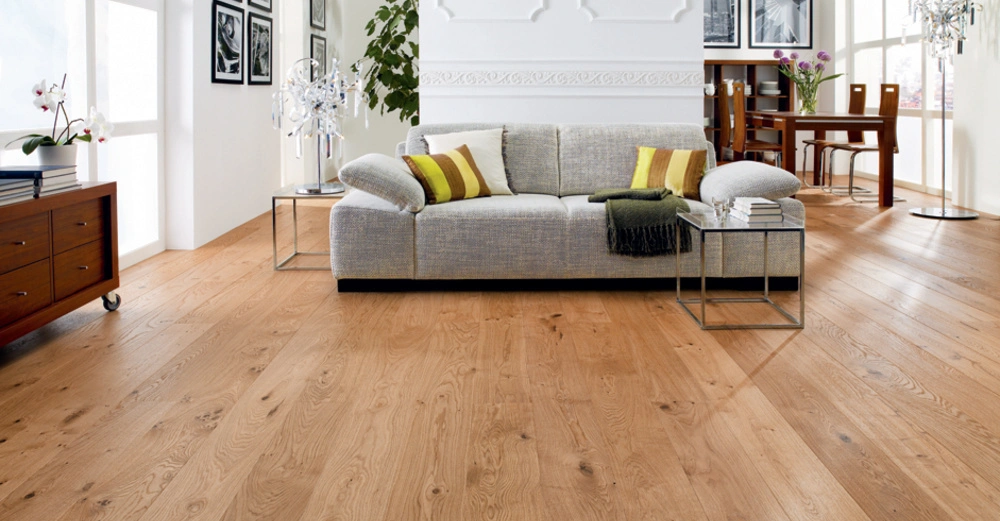 Graues Sofa mit grünen und gelben Kissen steht in einem hellen Wohnzimmer vor einem Tisch auf einem Holzfußboden; im Hintergrund ein Essbereich mit Holztisch und Stühlen sowie Wandbilder.