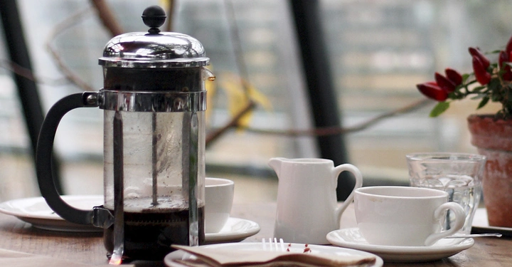 Eine Pressstempelkanne steht auf einem Holztisch neben Tassen Sahnekännchen und Teller in einem Café-Hintergrund mit Topfpflanze und unscharfer Hintergrundansicht durch Glasfenster.