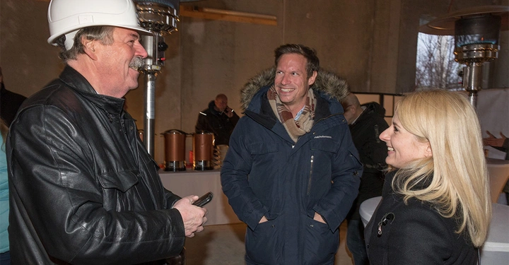 Galerie-Bild 4: Drei Personen stehen und unterhalten sich in einem Innenraum einer Baustelle. Der Mann links trägt einen Bauhelm hält ein Smartphone und lacht während die beiden anderen lachen und zuhören.