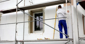Arbeiter in blitzblauer Arbeitshose bringt weiße Dämmplatten an der Fassade eines Einfamilienhauses an. Er steht auf einem Gerüst, neben im lehnt eine gelbe Wasserwaage an der Mauer.