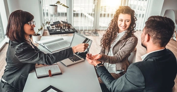 Eine Frau überreicht einem Paar einen Schlüssel ins Büro während sie am Tisch sitzen auf dem offene Bücher und ein Laptop liegen im Hintergrund ein Modellhaus.