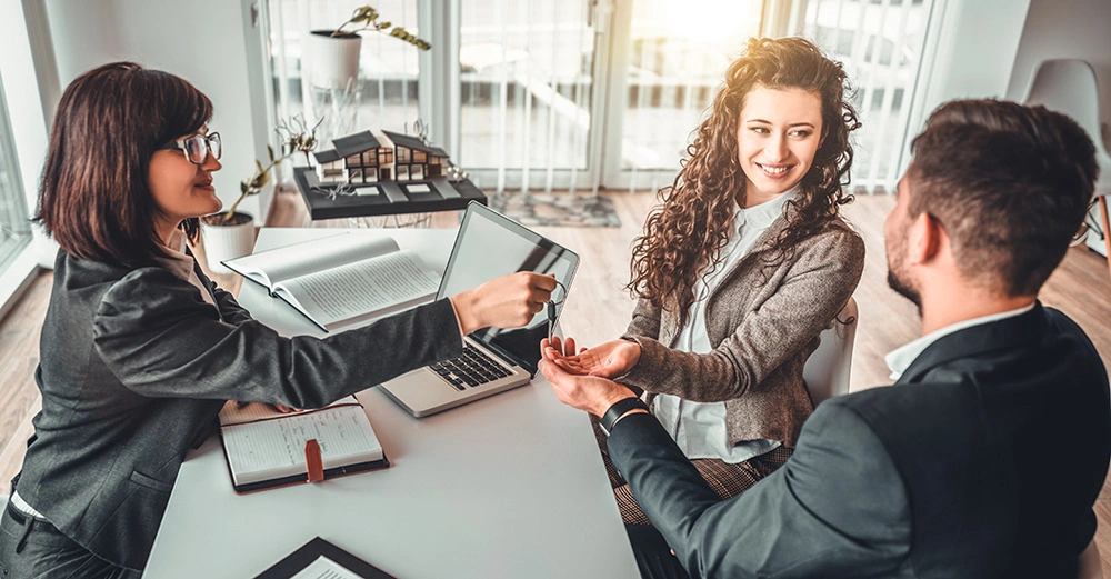 Eine Frau überreicht einem Paar einen Schlüssel ins Büro während sie am Tisch sitzen auf dem offene Bücher und ein Laptop liegen im Hintergrund ein Modellhaus.