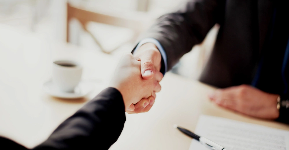 Zwei Personen schütteln sich die Hände in einem modernen Büro eine Tasse Kaffee und ein unterzeichneter Vertrag liegen auf dem Tisch