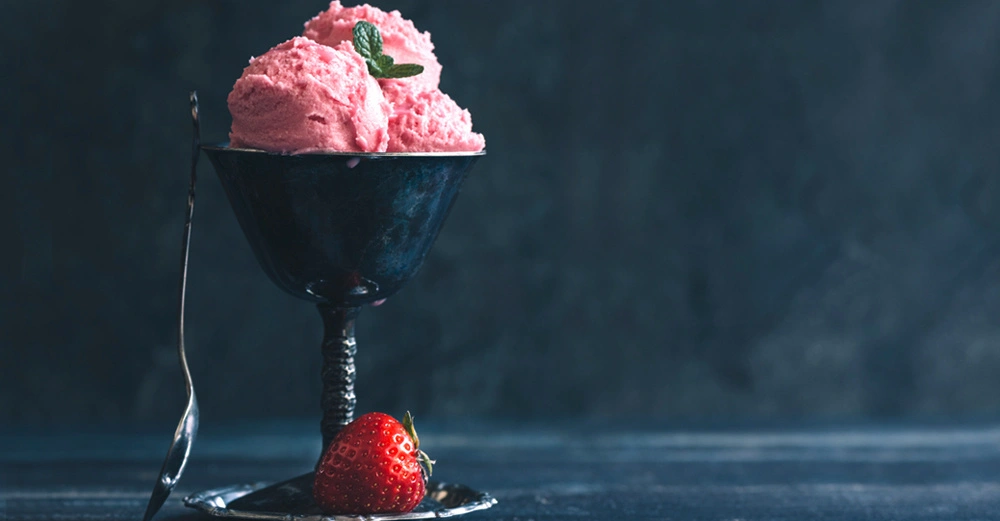 Rosa Eiscreme in einer silbernen Dessertschale mit einem Minzblatt dekoriert steht auf einem dunklen Tisch. Daneben liegt ein kleiner Löffel und eine frische Erdbeere. Hintergrund dunkel und unscharf.