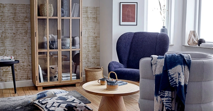Holzschrank steht neben blauem Sessel in einem Wohnzimmer Teekanne und Tasse auf einem runden Holztisch Teppiche und Kissen auf dem Holzboden Bilderrahmen an der Wand große Fenster enden