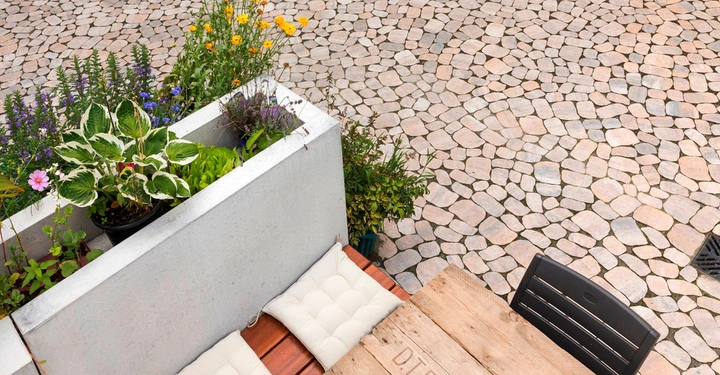 Weiße Sitzpolster auf einer Holzbänken daneben Pflanzgefäße mit bunten Blumen und grünen Pflanzen auf einer gepflasterten Terrasse im Freien