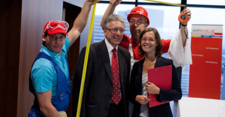 Galerie-Bild 41: Zwei Bauarbeiter halten ein Maßband über zwei lächelnde Personen in Anzügen in einem Bürogebäude die sich an einem Infostand mit dem Banner wohnnet.at befinden