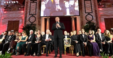 Mann steht und spricht in einem Ballsaal vor sitzendem Publikum in Abendkleidung. Im Hintergrund ist eine große Leinwand mit seinem Bild. Der Ballsaal hat Marmorsäulen und rote Beleuchtung.